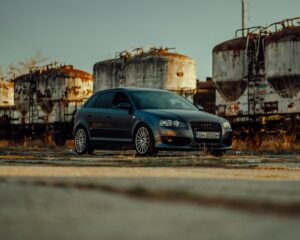 Gray car parked outdoors near rusted industrial tanks, creating a dramatic urban scene.